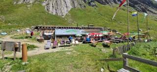 Alpenhütte mit Liegestühlen, Holzbänken und Sonnenschirmen auf einer Wiese vor einer felsigen Berglandschaft; Menschen sitzen und genießen das Wetter, im Vordergrund wehen mehrere Fahnen.