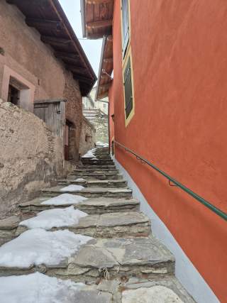 Steinige Außentreppe mit Schneeflecken zwischen zwei Gebäuden, eines davon mit roter Fassade und grünem Fensterladen. Ein grünes Handlauf entlang der roten Hauswand.