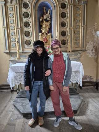 Zwei lächelnde Frauen stehen Arm in Arm vor einem Altar mit einer Statue der Madonna und Kind in einem historischen Kirchenraum.