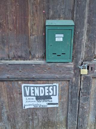 Alte Holztür mit grünem Briefkasten und handgeschriebenem Verkaufsschild „VENDESI“ mit Kontaktnummern, rechts daneben ein Vorhängeschloss.
