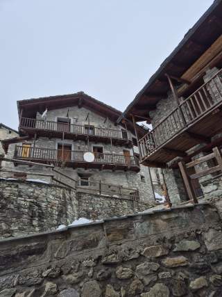 Blick nach oben auf traditionelle Steinhäuser mit Holzbalkonen und steilen Dächern, umrundet von Steinmauern und etwas Schnee.