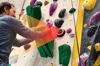 Mann klettert an einer bunten Kletterwand und berührt ein rundes Ziel mit leuchtenden roten Lichtern.