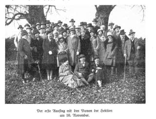 Schwarz-weiß-Gruppenfoto von etwa 40 Männern und Frauen in zeitgenössischer Kleidung und Hüten, aufgenommen im Freien vor zwei großen Bäumen. Eine Gruppe sitzt und der Großteil steht, alle blicken zur Kamera.