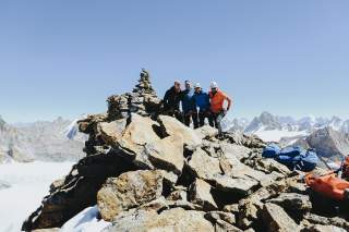 Vier Bergsteiger an einem hochalpinen Gipfel. Große Steinplatten sind zu sehen, im Hintergrund andere Gipfel und ein Gletscher.