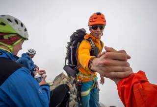 Ein Mann mit Helm reicht anderer Person die Hand auf den letzten Metern zum Gipfel