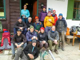 Gruppenfoto von 13 Personen, die vor einer Berghütte lächelnd zusammen sitzen und stehen, einige halten Tassen oder Becher in der Hand. Im Hintergrund sind Rucksäcke und Outdoor-Ausrüstung sichtbar.