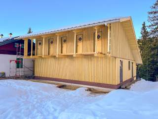 Ein Hausanbau aus Holz mit fünf Türen, die an der Außenwand auf einen noch unfertigen Laubengang führen, davor Schnee auf dem Boden.