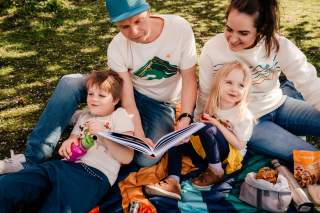 Familie (Mann, Frau, Junge, Mädchen) sitzen auf einer Picknickdecke auf einer Wiese und lesen gemeinsam ein Buch
