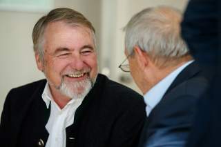 Zwei Männer unterhalten sich, einer von ihnen lächelt herzlich. Der lächelnde Mann trägt einen schwarzen Anzug und ein weißes Hemd, der andere eine Brille er sieht nicht in die Kamera.