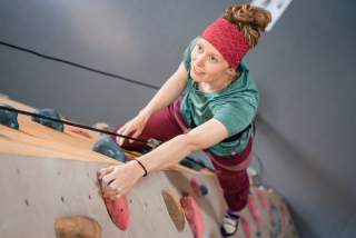 Frau mit rotem Stirnband klettert an einer Indoor-Kletterwand und blickt nach oben.