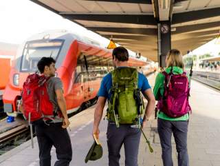 Drei Personen mit bunten Wanderrucksäcken stehen auf einem Bahnsteig vor einem roten Zug.