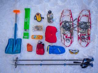 Ausrüstung für eine Winterwanderung, darunter Schneeschuhe, Teleskopstöcke, Schaufel, Lawinensonde, Lawinenpiepser, Thermosflasche, Sonnencreme, Mütze, Handschuhe, Gamaschen, Sonnenbrille und ein roter Packsack, auf Schnee ausgebreitet.