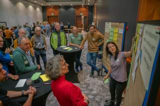 Eine Frau erklärt einer Gruppe von Menschen an zwei Pinnwänden mit Notizzetteln stehend Inhalte in einem Konferenzraum. Die Zuhörer hören aufmerksam zu und betrachten die Pinnwände.