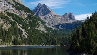 Blick auf einen kleinen, grünen See - rund herum Wald un im Hintergrund sieht man nah und deutlich einen steinernen Berg, der wie zwei Pyramidenspitzen als Gipfel hat.