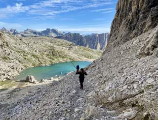 Zwei Menschen wandern in steinigem Gelände mit Bergsee und Hütte im Hintergrund