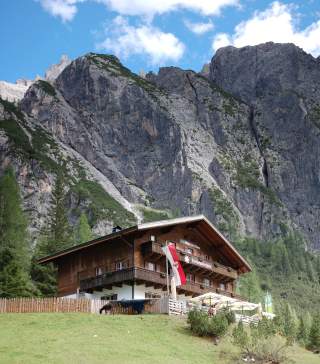 Hütte mit Balkon und Terrasse steht auf einer Wiese vor steilen, felsigen Bergen, eine rot-weiße Fahne weht davor.