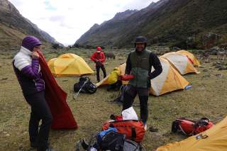 Gruppe von Wanderern in Outdoor-Bekleidung, die bei gelben Zelten in einem Bergtal campen und Ausrüstung sortieren. Im Hintergrund sind Berge und mehrere Esel zu sehen.