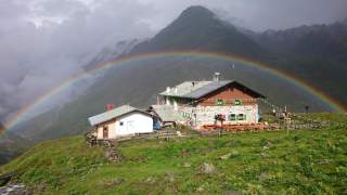 Hütte auf grüner Bergwiese vor einem nebelverhangenen Berg mit einem intensiven Regenbogen im Hintergrund.