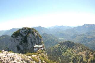 Berglandschaft mit einem markanten Felsen, auf dem eine Berghütte steht, umgeben von bewaldeten Hügeln und weiteren Bergen im Hintergrund.