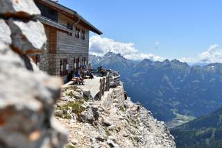 Berghütte mit Terrasse auf einem Felsvorsprung, Menschen sitzen draußen, im Hintergrund eine beeindruckende Alpenlandschaft unter blauem Himmel.