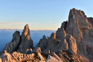 Raues, felsiges Bergmassiv im Sonnenuntergang, mit Wolkenmeer im Hintergrund und blauem Himmel.