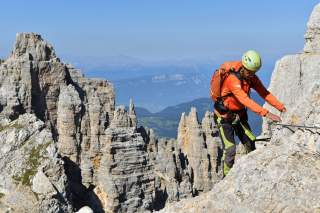Ein Bergsteiger mit Helm und orangefarbener Jacke klettert gesichert an einem Drahtseil entlang einer felsigen Bergwand, im Hintergrund steile Felsformationen und ein weites Tal.