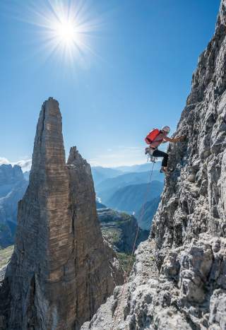 Ein Bergsteiger mit Helm und rotem Rucksack klettert an einer steilen Felswand, im Hintergrund ragt eine markante Felsspitze in die Höhe, die Sonne scheint am klaren Himmel.