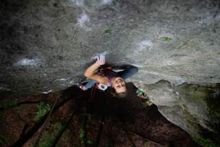 Junge Frau klettert an einer steilen Felswand, schaut lachend nach oben, gesichert mit Kletterausrüstung.