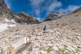 Wanderer mit Rucksack steigt auf felsigem, steinigem Bergpfad unter blauem Himmel, ein Wegzeichen ist auf einem großen Stein im Vordergrund zu sehen.