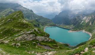 Berglandschaft mit grün bewachsenen Felsen, einem türkisfarbenen Stausee und bewölktem Himmel. Im Hintergrund sind hohe, teils schneebedeckte Berge zu sehen.