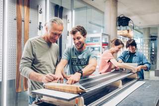 Vier Personen arbeiten gemeinsam an einem langen Ski, vermutlich bei der Montage oder Reparatur, in einer Werkstatt mit Ski im Hintergrund. Alle wirken konzentriert und teilen freundliche Stimmung.