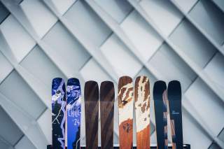 Mehrere Paar Skier mit unterschiedlichen Designs und Farben stehen nebeneinander vor einer geometrisch gemusterten, hellgrauen Wand.