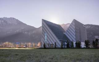 Modernes, dreieckiges Gebäude mit Metallfassade vor bewaldeten Bergen im Sonnenlicht.