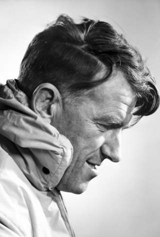 Schwarz-weiß-Profilporträt eines Mannes mit nach vorne geneigtem Kopf, der eine dicke Winterjacke trägt und lächelt.