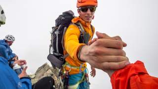 Bergsteiger in bunter Ausrüstung reicht helfend die Hand an eine weitere Person, im Hintergrund sitzen weitere Bergsteiger mit Helmen und Ausrüstung.