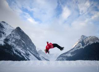 Eine Person in rotem Pullover macht rückwärts einen Sprung im Schnee vor einer Kulisse schneebedeckter Berge und blauem Himmel mit Wolken.