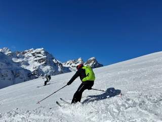 Zwei Skifahrer fahren auf einer schneebedeckten Piste bergab, im Hintergrund sind schneebedeckte Berge und blauer Himmel zu sehen.