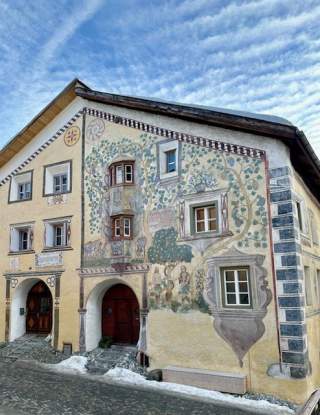 Fassadenansicht eines historischen Hauses mit bemalter Wandgestaltung, die Fensterrahmen, Pflanzen und Figuren zeigt. Vor dem Haus liegt eine gepflasterte Straße mit etwas Schnee.