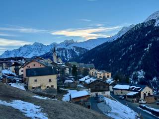 Bergdorf mit schneebedeckten Dächern vor einem Hintergrund aus hohen, schneebedeckten Bergen und blauem Himmel bei Sonnenuntergang.