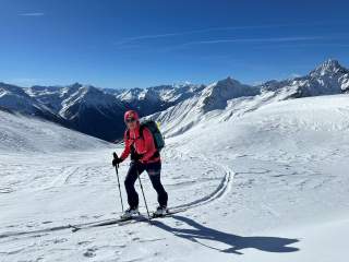 Person in roter Jacke und Sonnenbrille beim Skitourengehen in verschneiter Berglandschaft unter blauem Himmel.