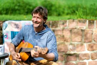 Ein lächelnder Mann sitzt draußen auf einem Stuhl und spielt Gitarre, im Hintergrund eine Steinmauer und grüne Wiese.