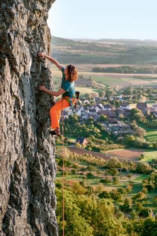 Eine Person klettert an einer steilen Felswand, im Hintergrund ist eine ländliche Landschaft mit Feldern und einem Dorf zu sehen.