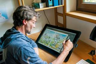 Ein Mann sitzt an einem Schreibtisch und zeichnet mit einem digitalen Stift einen Comic auf einem Grafiktablet.