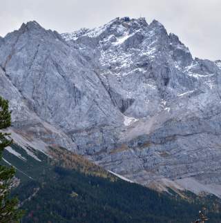 Große, schroffe Bergspitze mit etwas Schnee und einem Gebäude an der Gipfelkante, darunter dichte Nadelwälder. Der Himmel ist bewölkt.