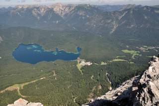 Blick von einem Felsen auf einen von Wald umgebenen, blaugrünen See mit Bergen im Hintergrund und einer kleinen Siedlung am Seeufer.