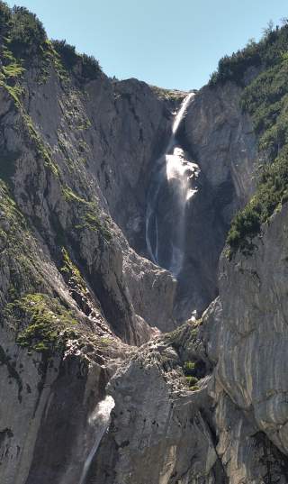 Ein Wasserfall stürzt zwischen steilen, felsigen Klippen mit vereinzeltem Grün herab, unter einem klaren blauen Himmel.