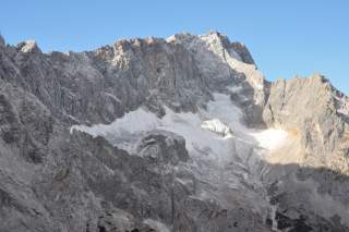 Felsige Berggipfel mit Schneefeldern vor klarem blauem Himmel, teilweise von Sonnenlicht und Schatten bedeckt.