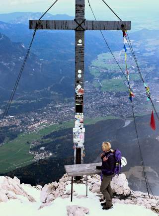 Wanderin steht auf schneebedecktem Berggipfel neben einem großen hölzernen Gipfelkreuz mit Aufklebern, im Hintergrund Panorama eines Tals mit Ortschaften und Bergen.