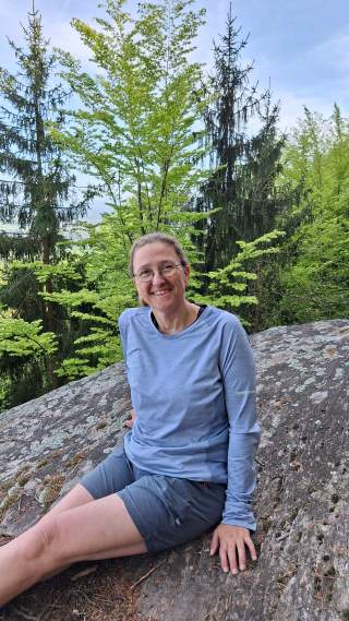Frau mit Brille, langärmligem hellblauem Oberteil und grauen Shorts sitzt auf einem großen Felsen, umgeben von grünen Bäumen in einem Wald.