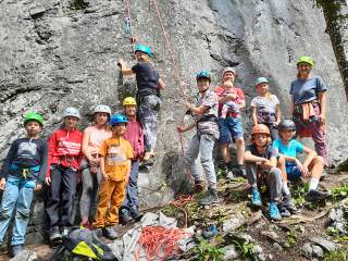 Eine Gruppe von Kindern und zwei Erwachsenen mit Kletterhelmen und Sicherungsausrüstung steht vor einer Felswand, eine Person klettert gerade. Am Boden liegen Kletterseile und Ausrüstung.